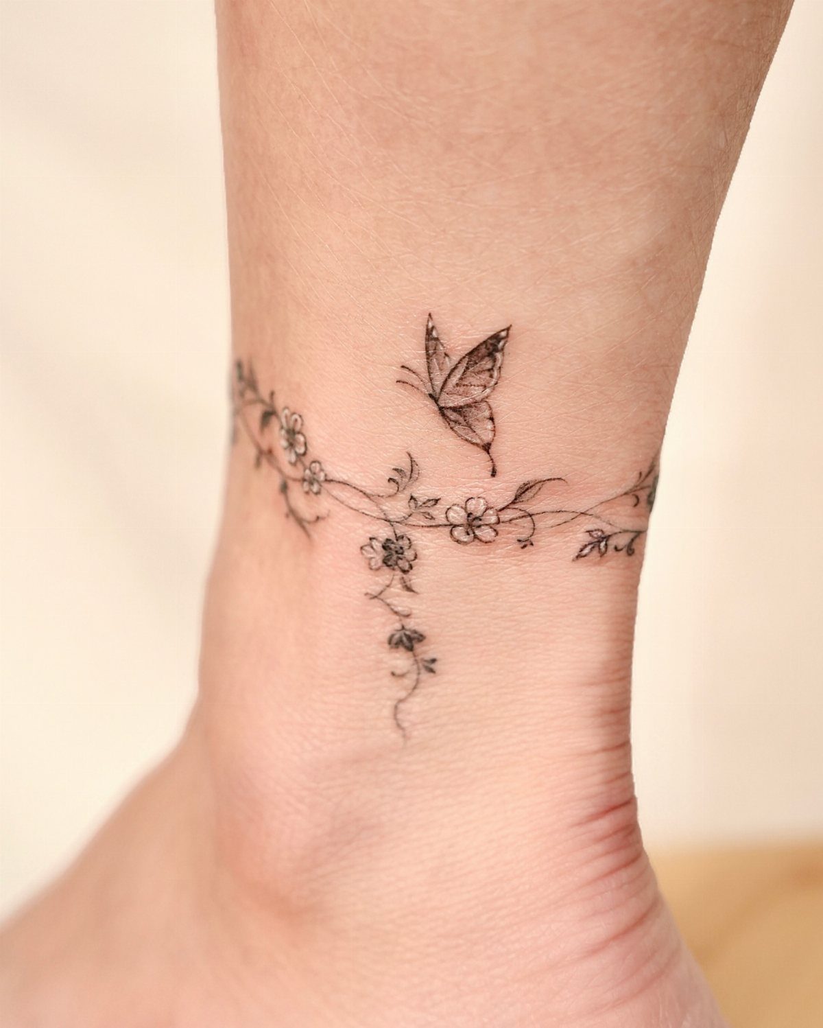Ankle tattoo featuring a delicate floral vine and a small butterfly in black ink.