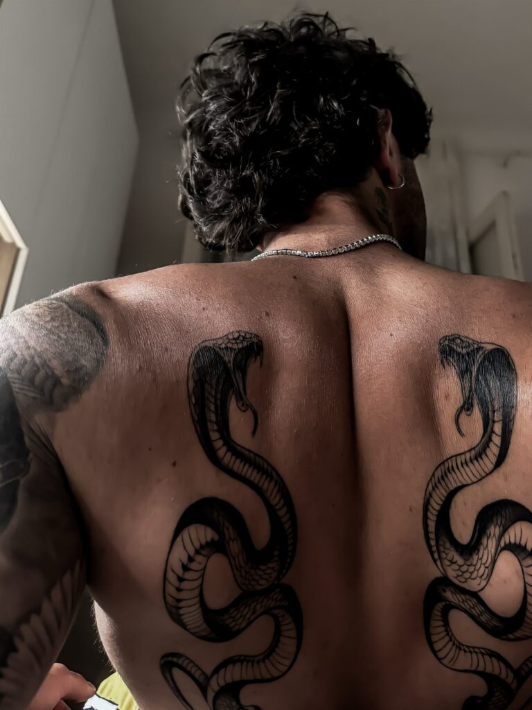 Man's back featuring two large black snake tattoos on his shoulder blades.