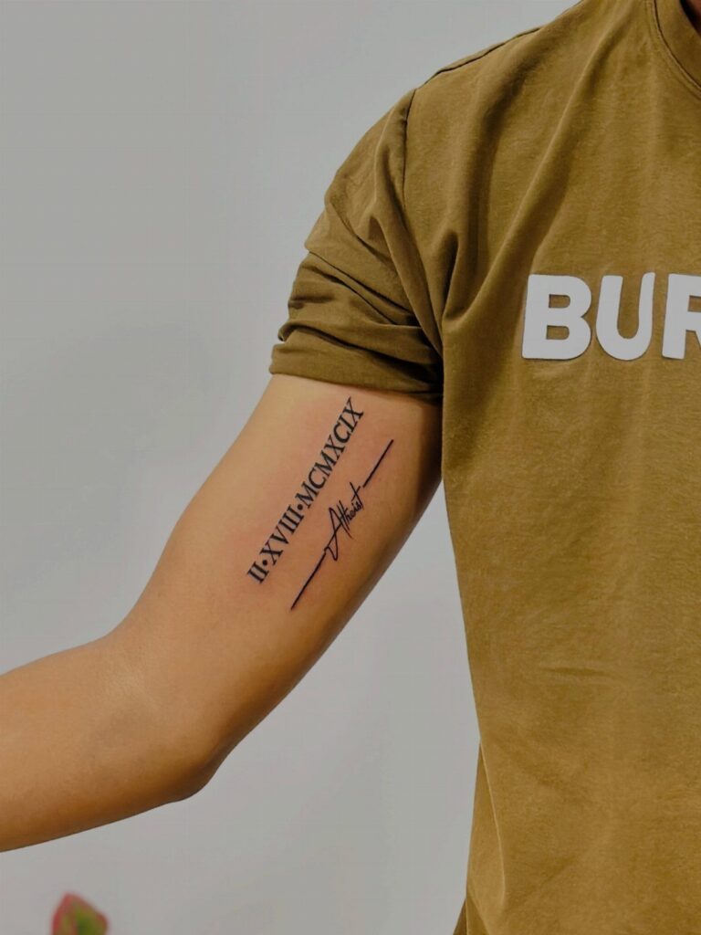 Bicep tattoo with Roman numerals and a signature on a man's arm.