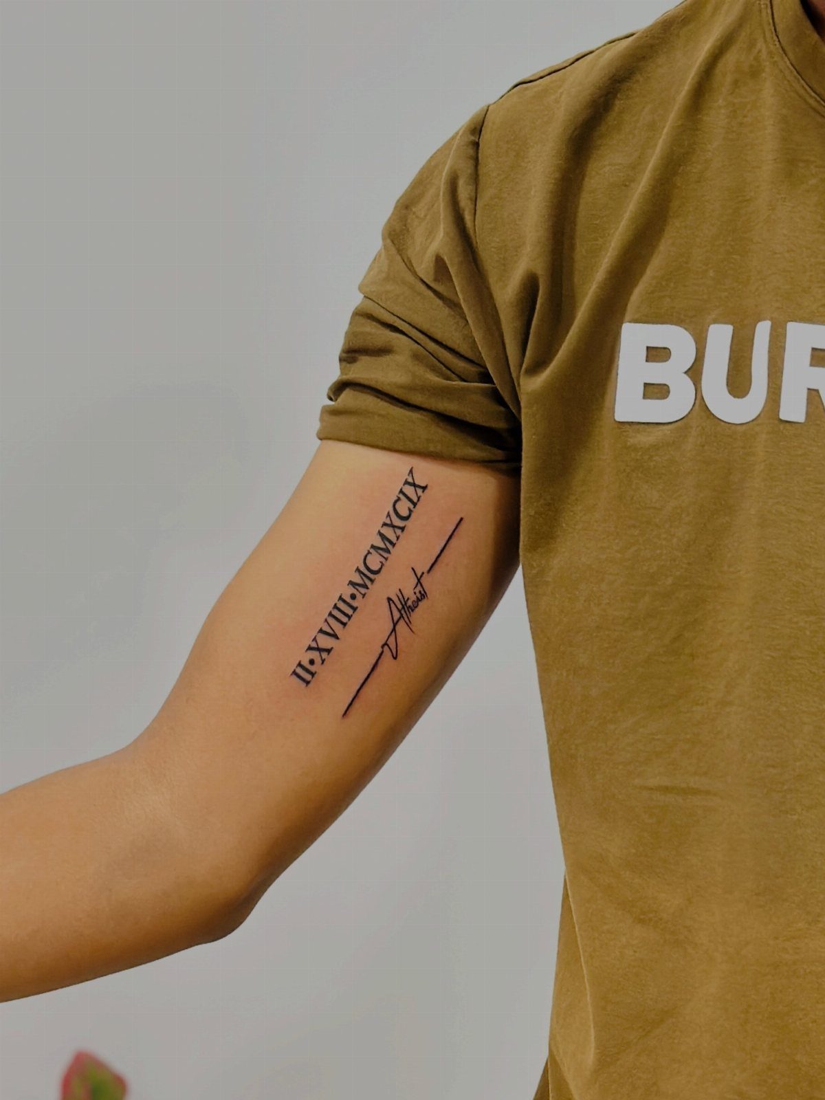 Bicep tattoo with Roman numerals and a signature on a man's arm.