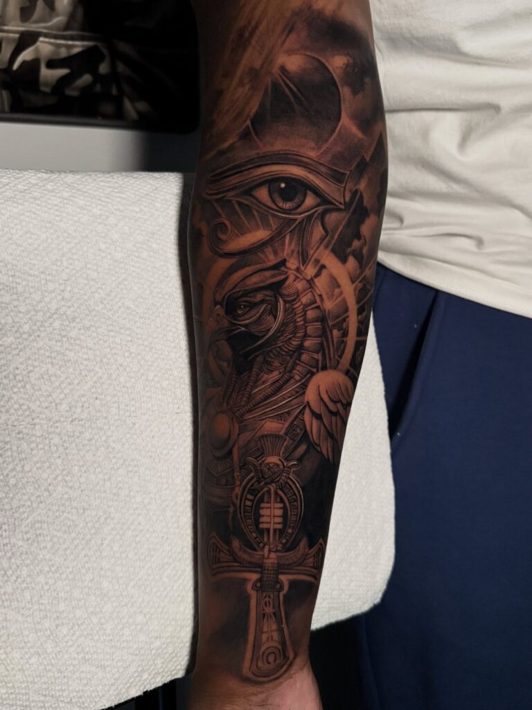 Forearm sleeve tattoo with black and grey Egyptian symbols including the Eye of Horus.