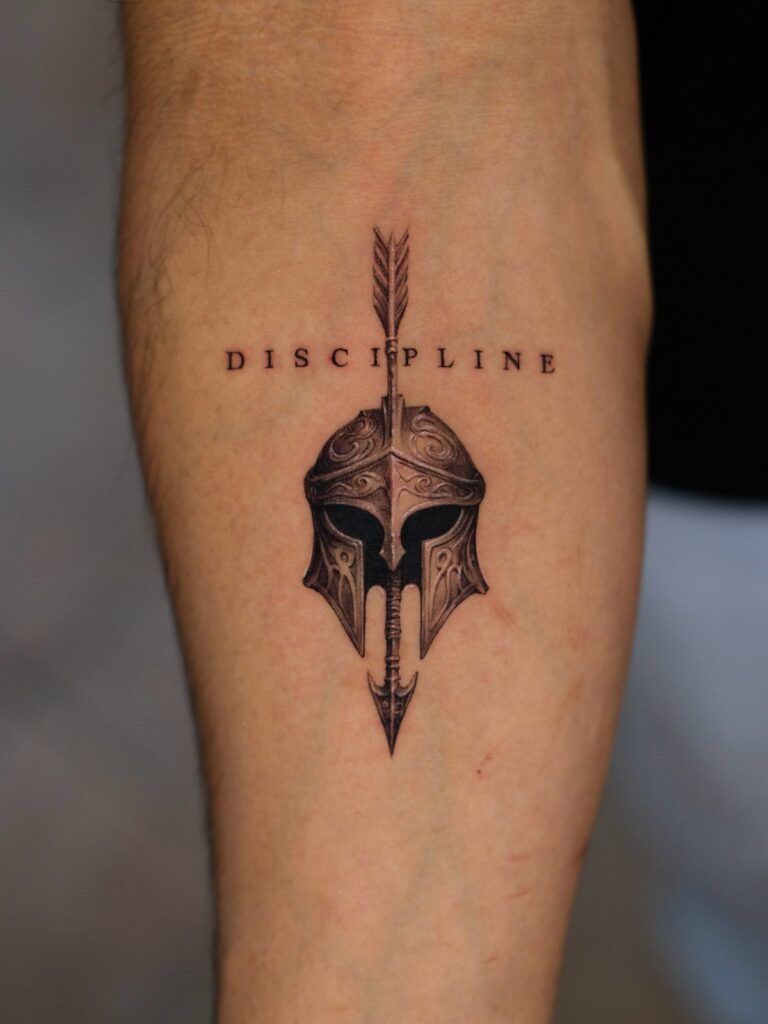 Spartan helmet and arrow tattoo with the word DISCIPLINE on a man's forearm