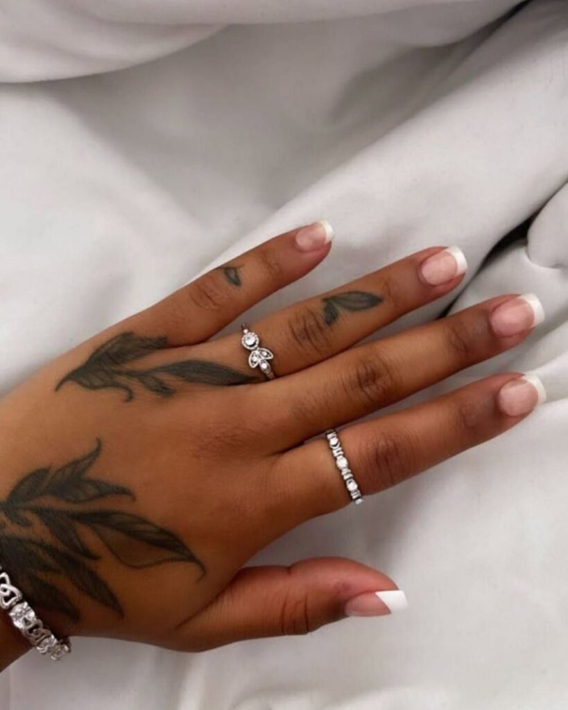 Hand with leaf tattoos, French tip nails, and silver diamond rings on white fabric.