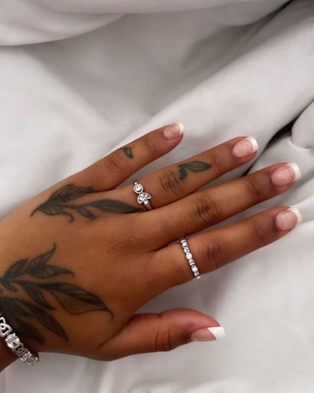 Hand with leaf tattoos, French tip nails, and silver diamond rings on white fabric.