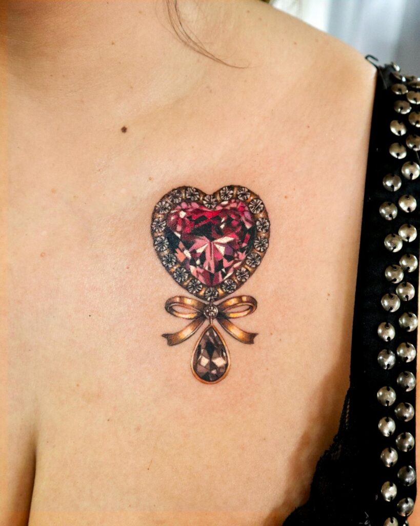 Heart-shaped jewel tattoo with a red gemstone, gold frame, and hanging bow.