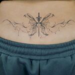 Sword and fairy wing tattoo on a person's lower back above grey sweatpants.