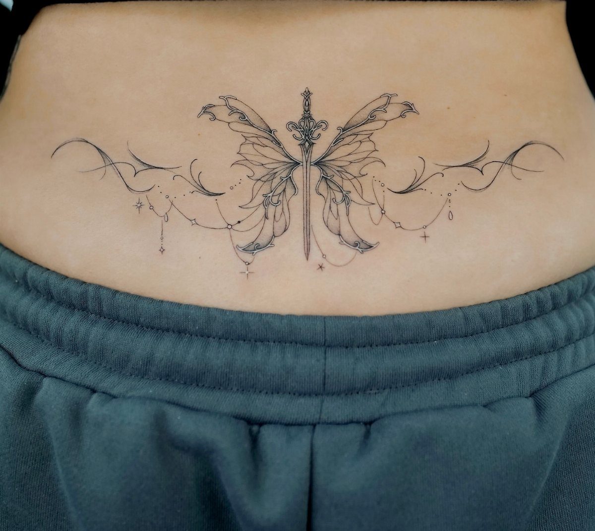 Sword and fairy wing tattoo on a person's lower back above grey sweatpants.