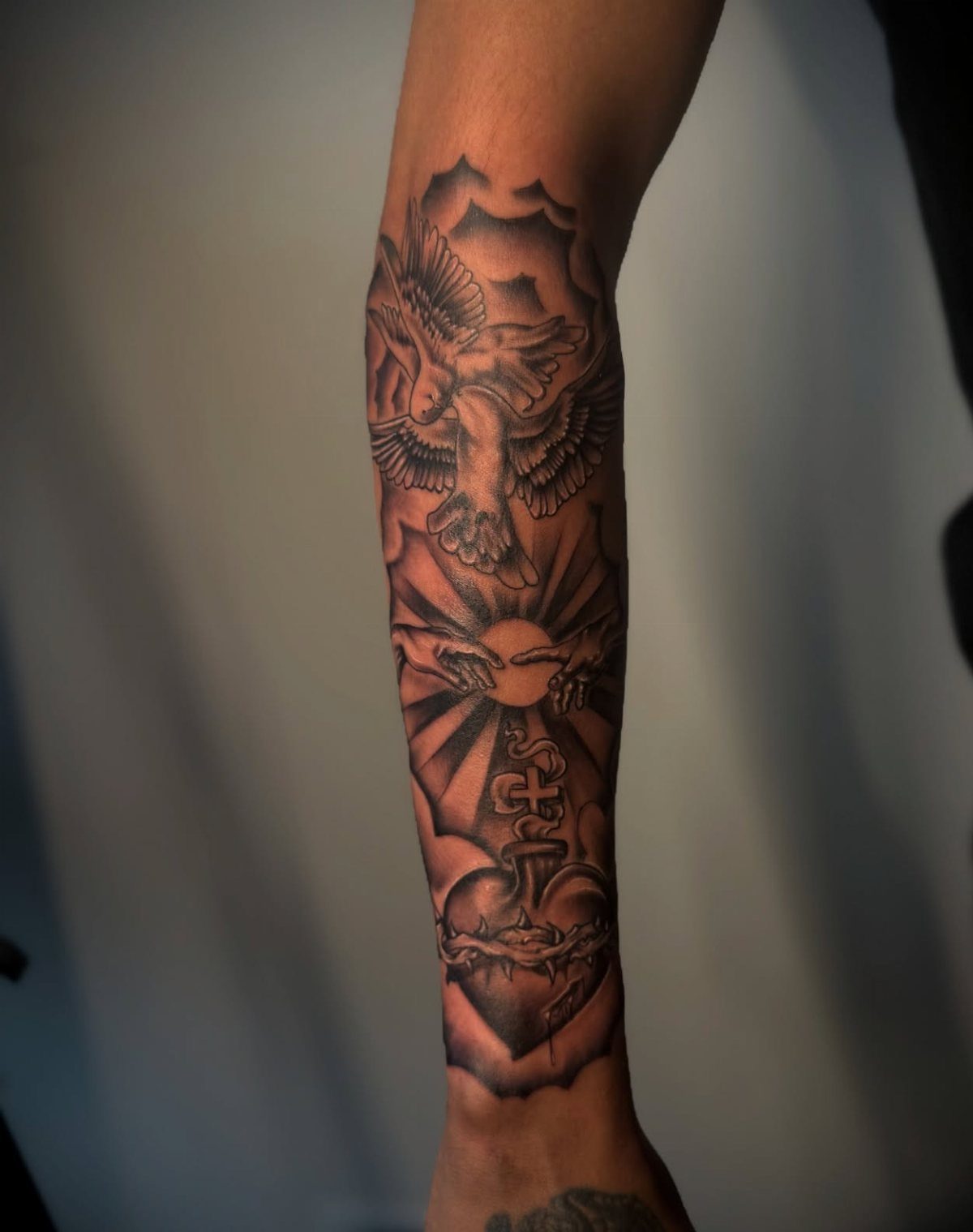 Forearm tattoo with a dove, hands, and sacred heart in black and grey ink.