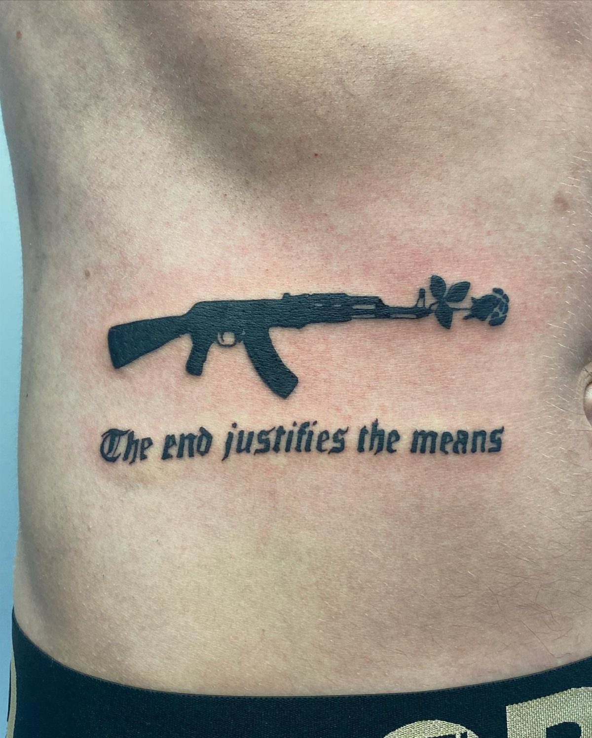 Rib tattoo of an AK-47 rifle with a rose and gothic text on skin.