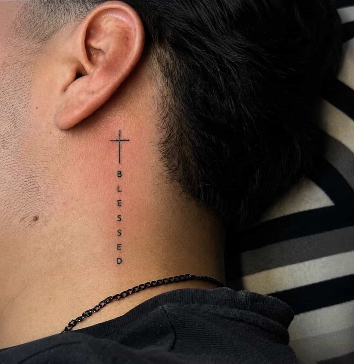 Side neck tattoo of a small cross and the word BLESSED in black ink.