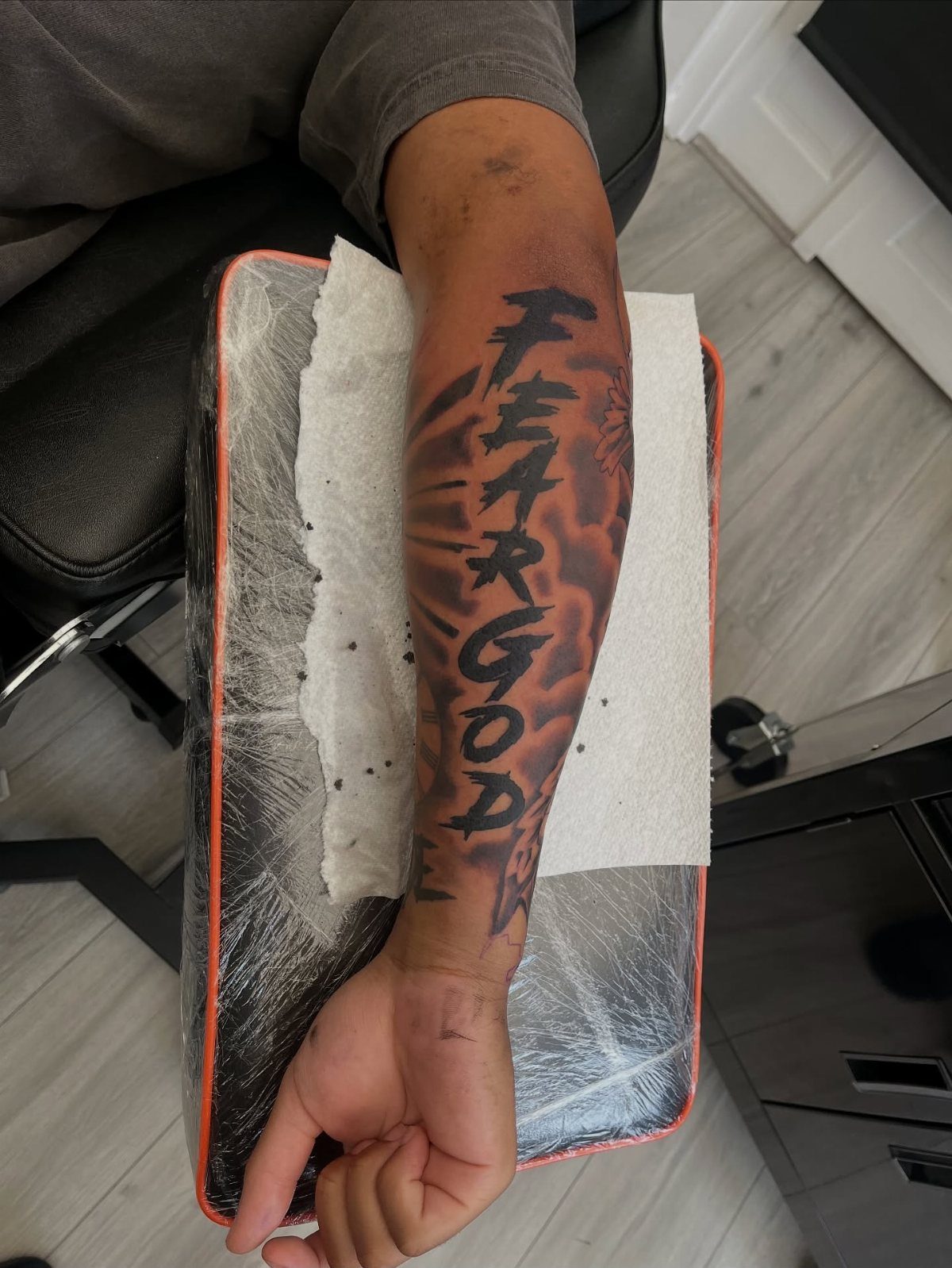 Man's forearm with a black ink tattoo reading FEAR GOD on an armrest.
