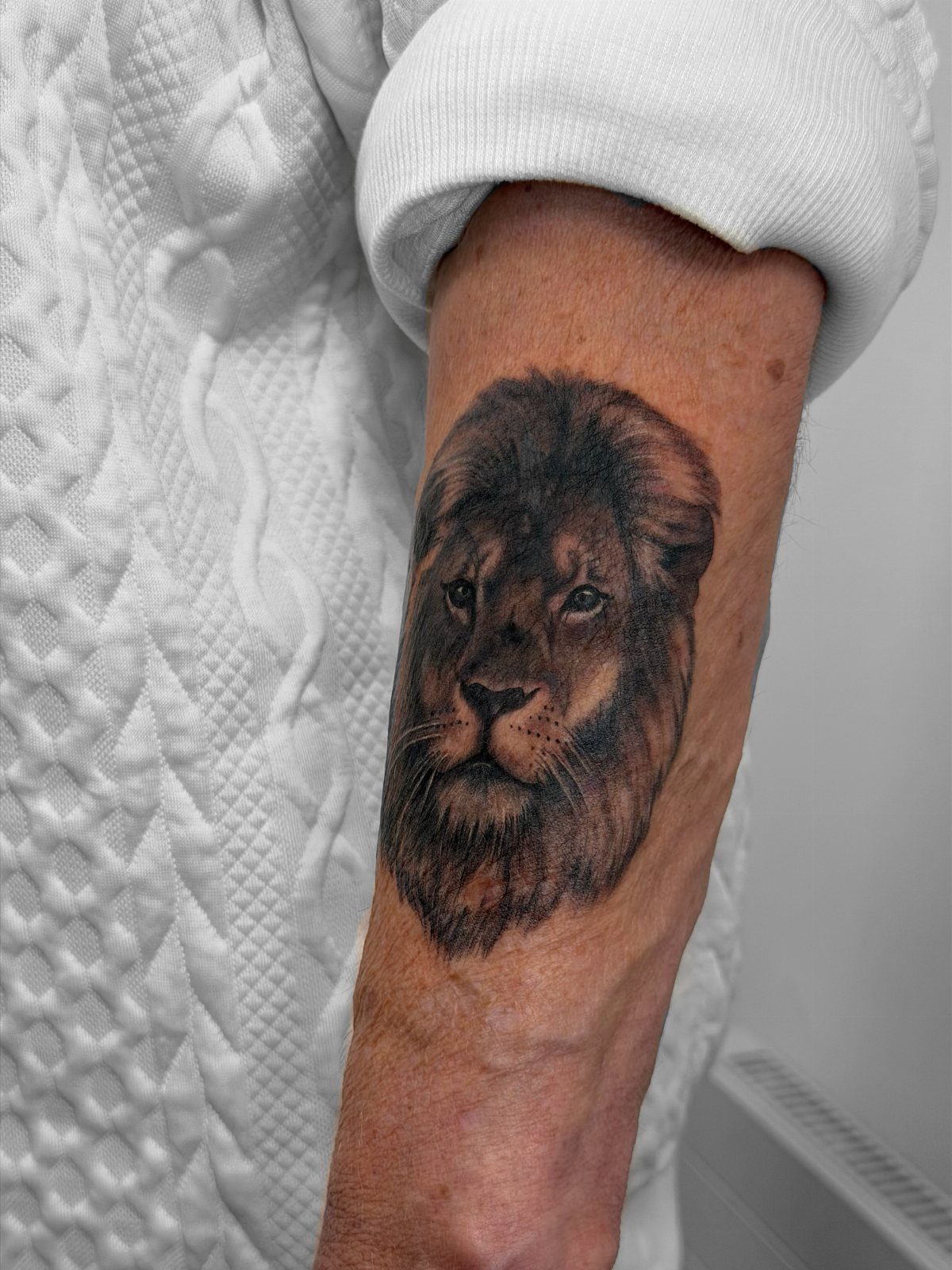 Lion head tattoo on the forearm of an older person wearing white clothing.
